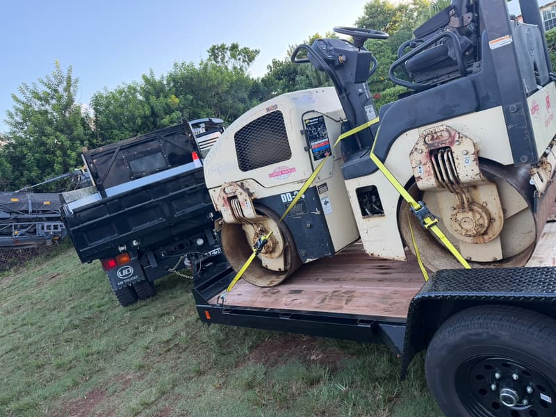 Quality Control tandem asphalt roller strapped to a trailer beside a dump truck